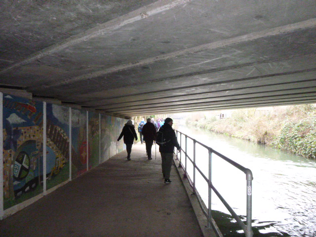 People walking under bridge beside canal, mural visible.